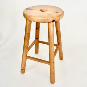 May include: A light-coloured wooden stool with a round seat and three legs. The stool has a simple design with a natural wood grain finish. The seat has a small, round indentation in the centre.