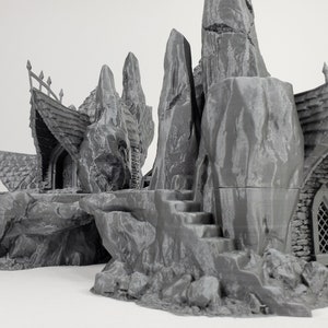May include: A 3D printed miniature of a stone building with a pointed roof and a small window. The building is surrounded by rocky terrain and appears to be perched on a cliff.