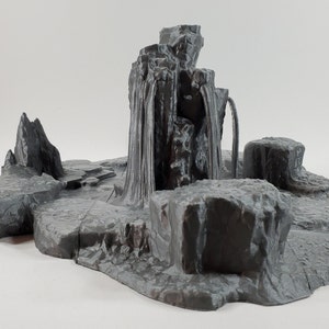May include: A gray miniature landscape with a waterfall feature. The scene includes rocky formations, a central waterfall, and a surrounding base. The design is detailed, with textured surfaces and a naturalistic aesthetic, suitable for tabletop gaming or model displays.