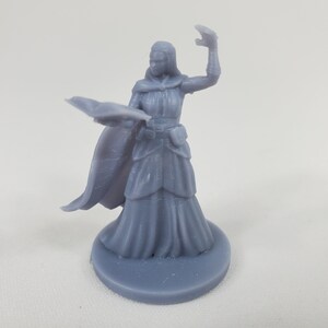 May include: A gray 3D printed miniature of a female figure wearing a long robe and a cloak. She is holding a book in her left hand and her right hand is raised in a gesture of casting a spell.