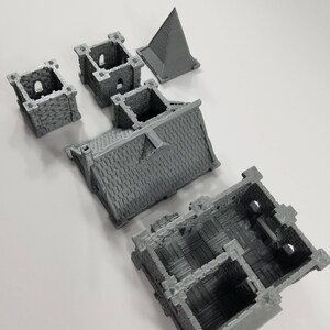 May include: A set of five gray 3D printed miniature building pieces. The pieces include a roof, a wall, a tower, and two smaller towers. The pieces are designed to be assembled into a small building.