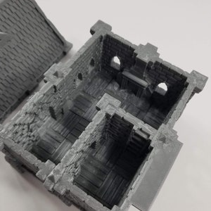May include: A gray 3D printed model of a small, multi-room building with a stone exterior and wooden floors. The building has a small, open doorway and several windows.