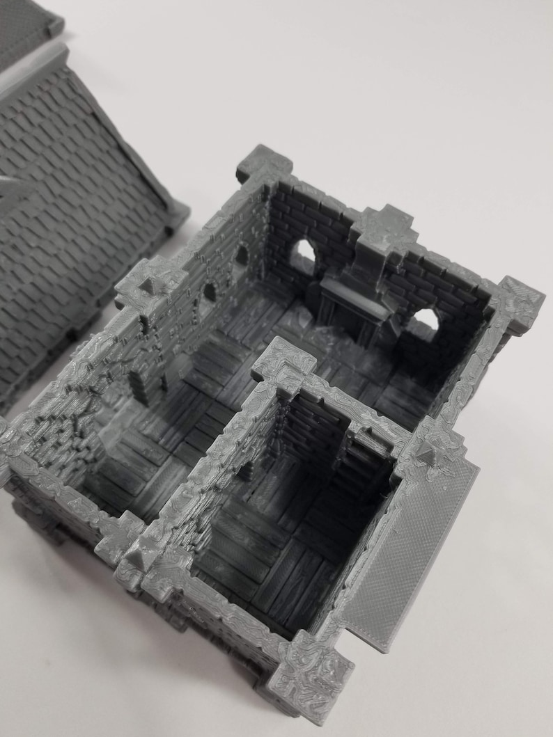 May include: A gray 3D printed model of a small, multi-room building with a stone exterior and wooden floors. The building has a small, open doorway and several windows.