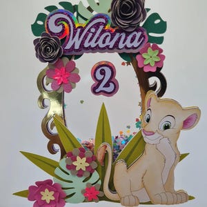 May include: A cake topper featuring a lion cub with a pink nose and green eyes, sitting on a bed of green leaves and pink flowers. The topper also includes a gold frame with the name "Wilona" and the number "2" in purple glitter.