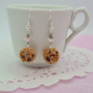 Peut inclure: Une paire de boucles d'oreilles pendantes argentées avec un charme de cookie aux pépites de chocolat et un charme de cœur argenté. Les boucles d'oreilles sont présentées sur une tasse blanche et une dentelle rose.