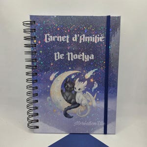 Peut inclure: Cahier à spirales bleu avec l'inscription "Carnet d'Amitié De Noëlya" et une illustration de dragon. La couverture présente un fond scintillant étoilé et une fermeture élastique bleue.
