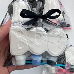 May include: A white ceramic "BOO" shaped craft kit, packaged in clear cellophane with a black ribbon bow. The kit includes small paint pots in white, blue, and black, and a small paintbrush. The background shows other similar kits.