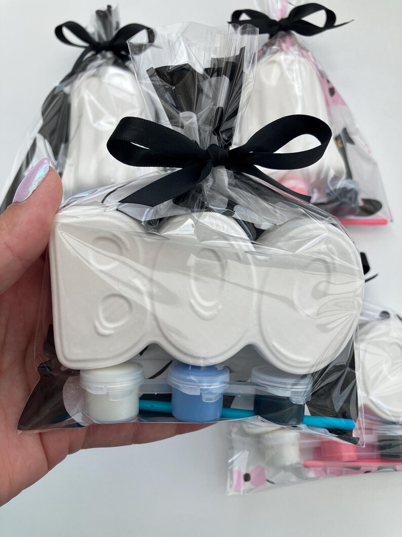 May include: A white ceramic "BOO" shaped craft kit, packaged in clear cellophane with a black ribbon bow. The kit includes small paint pots in white, blue, and black, and a small paintbrush. The background shows other similar kits.