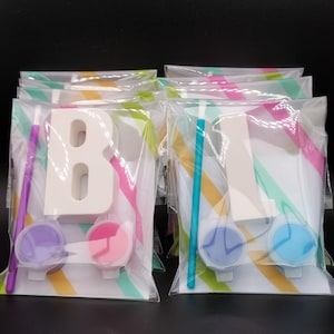 Può includere: Un set di 10 lettere di legno da dipingere, ogni lettera viene fornita con un piccolo pennello e tre piccoli barattoli di vernice. Le lettere sono in sacchetti di plastica trasparente con uno sfondo a strisce colorate.