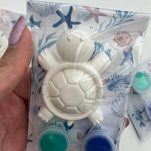 May include: A white turtle-shaped craft kit in a clear plastic bag. The kit includes a turtle figurine and two small paint pots, one green and one blue. The bag has a patterned design with seashells and starfish.