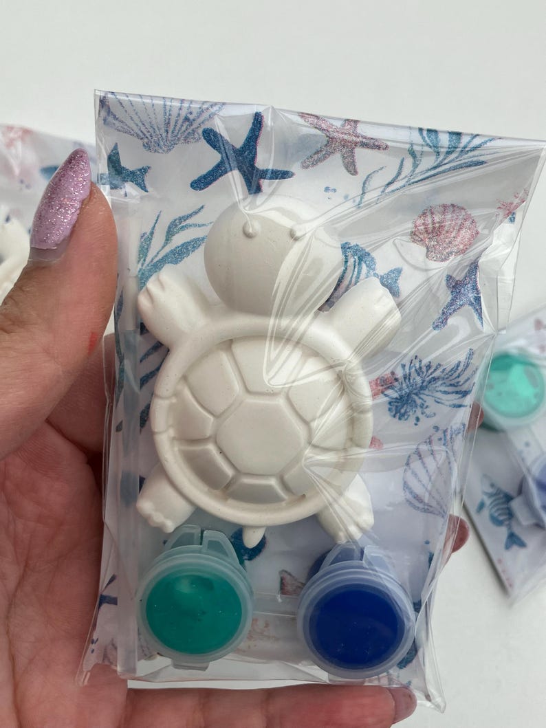 May include: A white turtle-shaped craft kit in a clear plastic bag. The kit includes a turtle figurine and two small paint pots, one green and one blue. The bag has a patterned design with seashells and starfish.