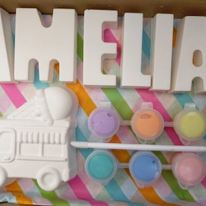 Pode incluir: Um kit de artesanato com o nome "AMELIA" em letras de cerâmica branca, um caminhão de sorvete de cerâmica branca e seis pequenos potes de tinta em roxo, laranja, amarelo, turquesa, azul e rosa.