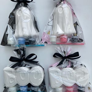 May include: Four Halloween-themed craft kits in clear cellophane bags tied with black bows. Two kits feature white ghost shapes, and two say "BOO" in white letters. Each kit includes paint pots and a paintbrush.