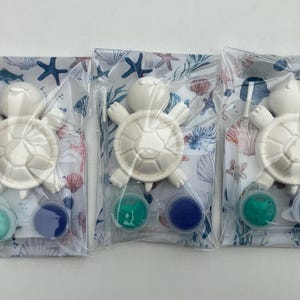 May include: Three craft kits, each containing a white turtle figurine, paint, and a brush. The kits include a turtle, two paint colours, and a small brush, all packaged in a clear bag with a sea-themed design.
