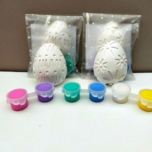 May include: Two white ceramic Easter eggs, one with a wavy pattern and the other with a flower pattern, are packaged in clear plastic bags. Six small paint pots in pink, purple, green, blue, white, and yellow are arranged in a row.