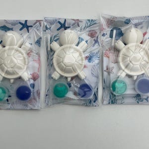 May include: Three clear plastic bags, each with a white turtle-shaped item and two small paint pots in teal and blue. The bags have a seashell and starfish pattern. The turtle has a shell design.