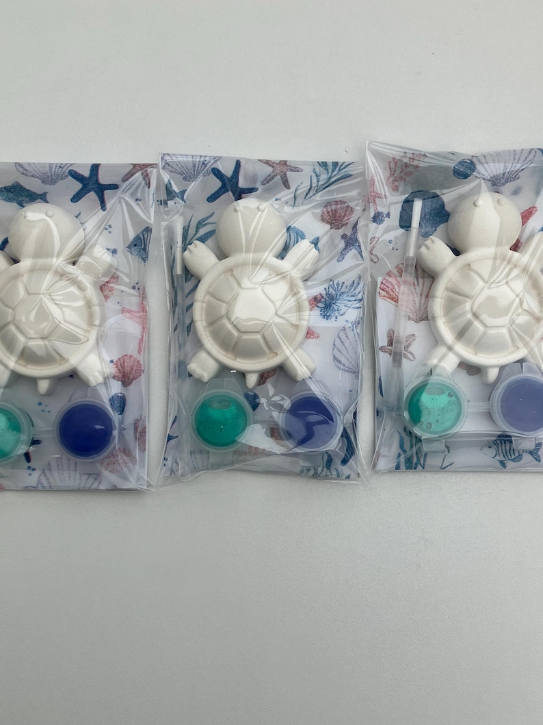 May include: Three clear plastic bags, each with a white turtle-shaped item and two small paint pots in teal and blue. The bags have a seashell and starfish pattern. The turtle has a shell design.