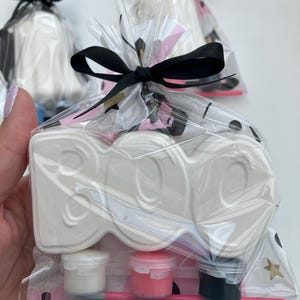 May include: A white ceramic "BOO" shaped craft kit, packaged in a clear cellophane bag and tied with a black ribbon. The kit includes small paint pots in white, pink, and black, along with a pink paintbrush. The background is white.