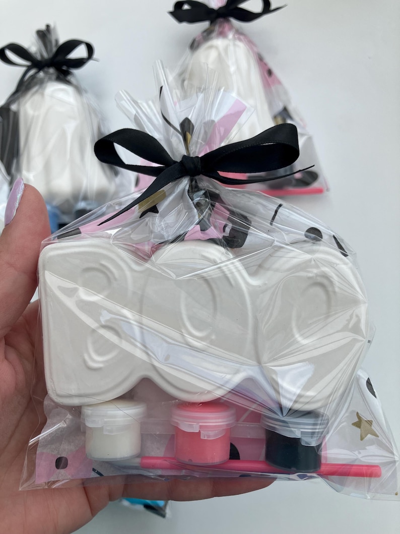 May include: A white ceramic "BOO" shaped craft kit, packaged in a clear cellophane bag and tied with a black ribbon. The kit includes small paint pots in white, pink, and black, along with a pink paintbrush. The background is white.
