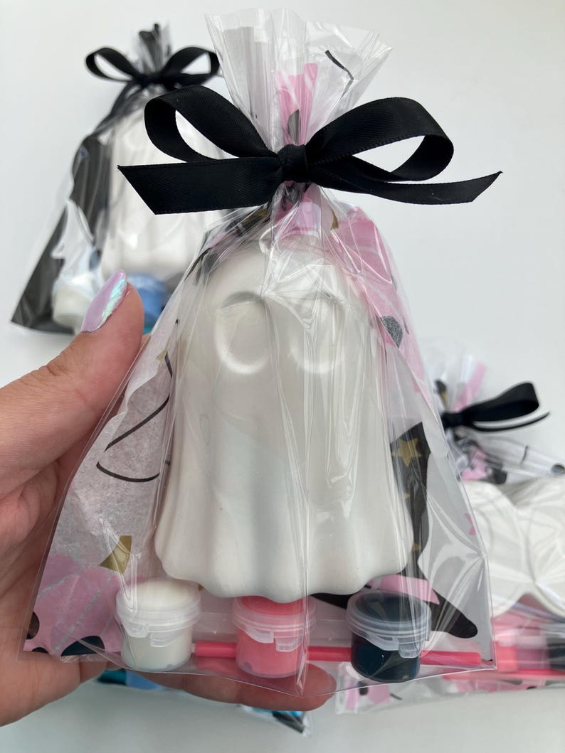 May include: White ghost-shaped craft kit in a clear cellophane bag, tied with a black satin ribbon. The kit includes small paint pots in white, pink, and dark blue, with paintbrushes. The background shows other similar kits.