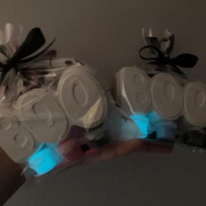 May include: Two clear cellophane bags tied with black ribbons. Each bag contains white, block-lettered "BOO" decorations and glowing blue accents. The bags are held against a dark background.