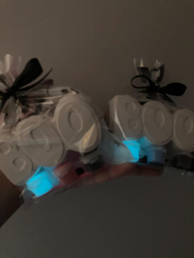 May include: Two clear cellophane bags tied with black ribbons. Each bag contains white, block-lettered "BOO" decorations and glowing blue accents. The bags are held against a dark background.