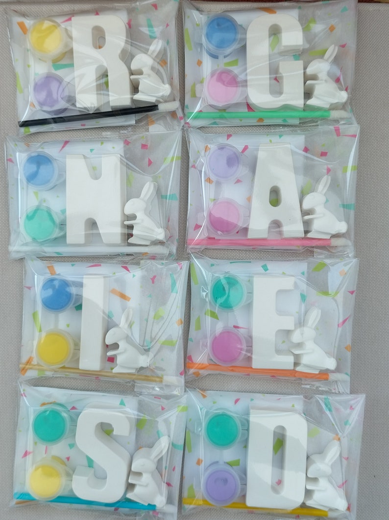May include: Eight individual clear plastic bags with white letters, paint pots, and small white bunny figurines. The letters spell out the word 'RAINBOW'. Each bag contains a different color paint pot: yellow, blue, purple, green, pink, blue, yellow, and green. Each bag also contains a small white bunny figurine. The bags are on a white and pink confetti background.