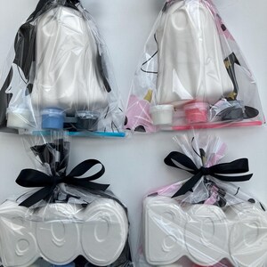 May include: Four Halloween-themed craft kits. Two kits feature white ghost figures, and two feature the word "BOO" in white. Each kit is packaged in a clear bag and tied with a black bow. Paint pots and brushes are included.