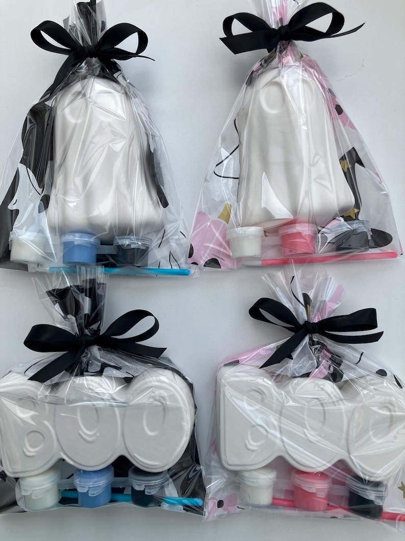 May include: Four Halloween-themed craft kits. Two kits feature white ghost figures, and two feature the word "BOO" in white. Each kit is packaged in a clear bag and tied with a black bow. Paint pots and brushes are included.