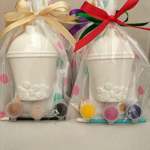 May include: Two ceramic bubble tea cups in clear plastic bags with colourful polka dots. Each bag has a ribbon tied around it and includes paint pots, a paintbrush, and glitter.