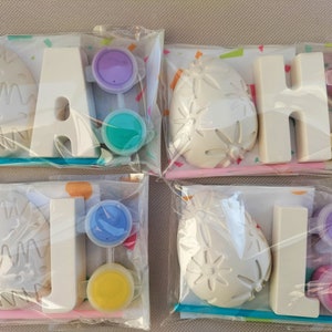 Può includere: Quattro sacchetti di plastica trasparente, ciascuno contenente un kit fai-da-te per dipingere. Ogni kit include una lettera bianca, una forma decorativa e vasetti di vernice di vari colori. Le lettere compongono la parola "JAHJ".