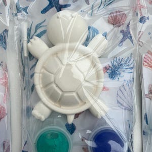 May include: A white ceramic turtle figurine with a segmented shell, packaged with two small paint pots of teal and blue. The packaging features a sea-themed pattern with starfish and seashells. The figurine is designed for painting.