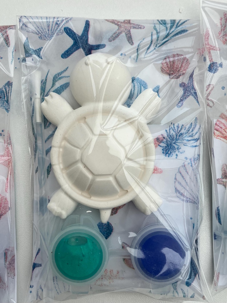 May include: A white ceramic turtle figurine with a segmented shell, packaged with two small paint pots of teal and blue. The packaging features a sea-themed pattern with starfish and seashells. The figurine is designed for painting.