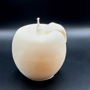 Peut inclure: Une bougie en forme de pomme de couleur cr&egrave;me, avec une m&egrave;che sur le dessus et un d&eacute;tail en forme de feuille sur le c&ocirc;t&eacute;. La bougie a une finition lisse et mate, sur fond sombre.