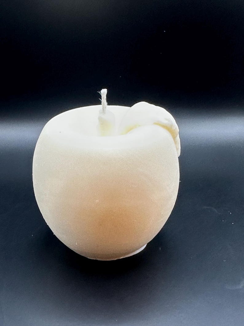 Peut inclure: Une bougie en forme de pomme de couleur cr&egrave;me, avec une m&egrave;che sur le dessus et un d&eacute;tail en forme de feuille sur le c&ocirc;t&eacute;. La bougie a une finition lisse et mate, sur fond sombre.