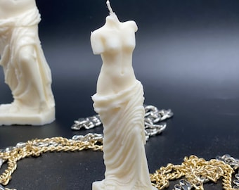 Op de afbeelding: Witte gebeeldhouwde kaars van het standbeeld van de Venus van Milo. De kaars staat op een zwart oppervlak met gouden en zilveren kettingen.