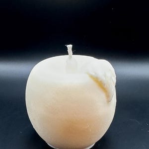 Peut inclure: Une bougie en forme de pomme, de couleur cr&egrave;me, avec une m&egrave;che blanche. La bougie a une forme lisse et arrondie avec une l&eacute;g&egrave;re texture et un petit d&eacute;tail de cire fondue sur le c&ocirc;t&eacute;. Le fond est noir uni.