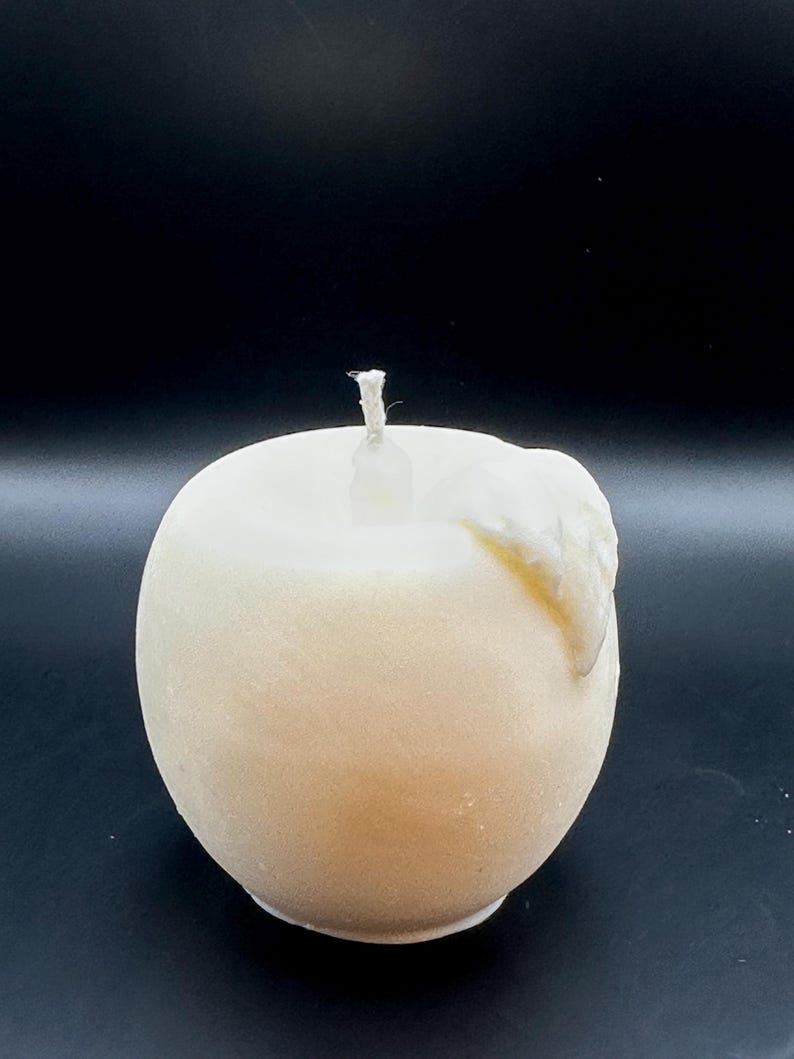 Peut inclure: Une bougie en forme de pomme, de couleur cr&egrave;me, avec une m&egrave;che blanche. La bougie a une forme lisse et arrondie avec une l&eacute;g&egrave;re texture et un petit d&eacute;tail de cire fondue sur le c&ocirc;t&eacute;. Le fond est noir uni.