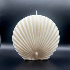 May include: A cream-colored, seashell-shaped candle with a textured surface. The candle has a central wick and a fan-like design, with vertical ridges radiating from a central point. The candle is set against a black background.