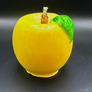 Peut inclure: Une bougie jaune en forme de pomme avec une feuille verte. La bougie a une m&egrave;che marron et est pos&eacute;e sur un fond noir. Article d&eacute;coratif.