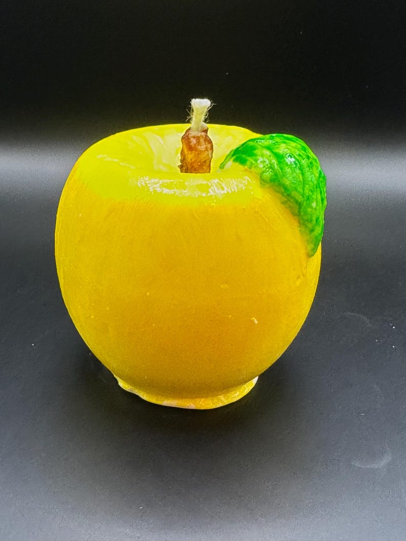 Peut inclure: Une bougie jaune en forme de pomme avec une feuille verte. La bougie a une m&egrave;che marron et est pos&eacute;e sur un fond noir. Article d&eacute;coratif.