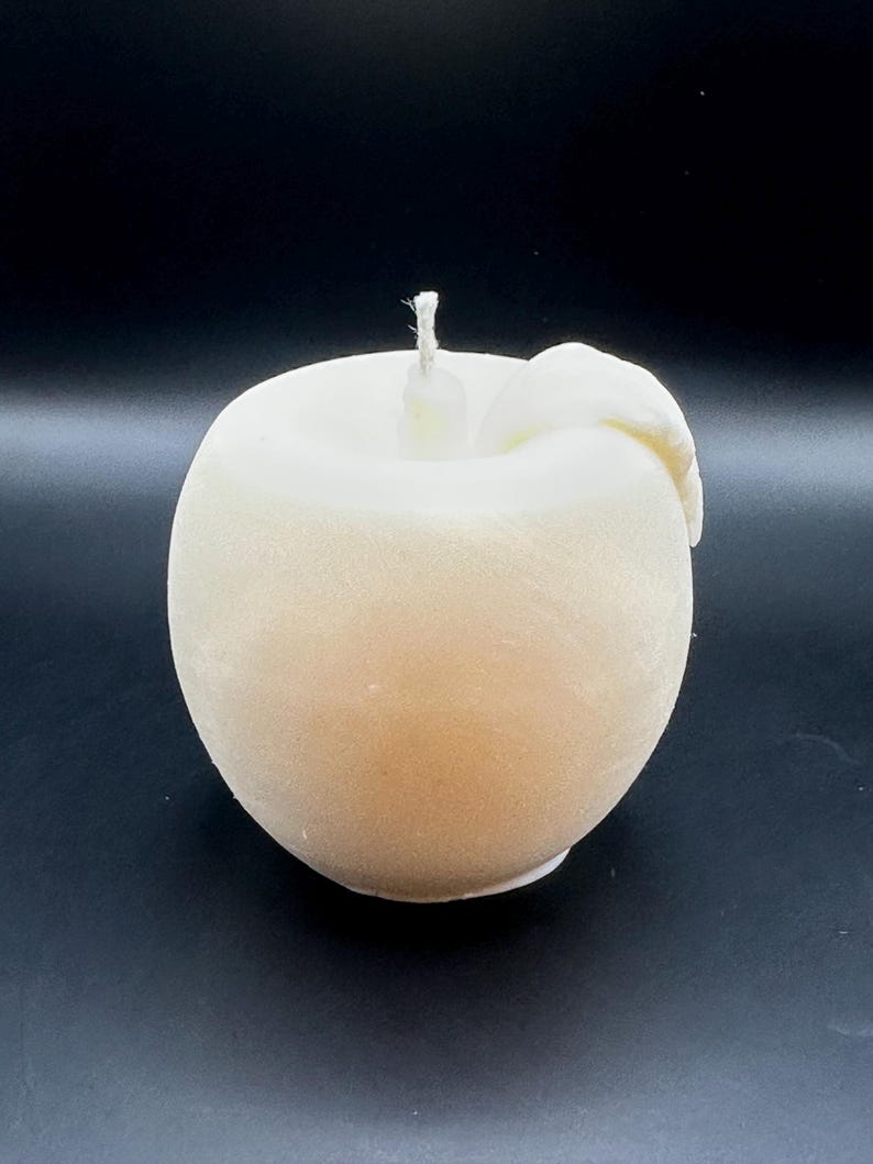 Peut inclure: Une bougie en forme de pomme de couleur cr&egrave;me, avec une m&egrave;che blanche et un d&eacute;tail en forme de feuille. La bougie a une forme lisse et arrondie, sur fond sombre. Cette bougie d&eacute;corative est con&ccedil;ue pour ressembler &agrave; une pomme fra&icirc;che.