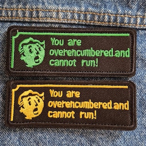 May include: Two embroidered patches with the text "You are overencumbered and cannot run!". One patch has a green border and character, the other has a yellow border and character. Both are on a dark brown background.