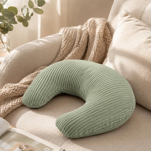 Könnte beinhalten: Ein halbmondförmiges Kissen aus salbeigrünem Cordstoff mit einer ausgeprägten gerippten Textur. Das Kissen hat ein geschwungenes Design und liegt auf einer gepolsterten Oberfläche in neutraler Farbe, was das weiche Material hervorhebt.