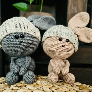 May include: Two handmade crochet dolls, one grey and one tan, each with a cream-coloured cap and black button eyes. The grey doll has a matching grey body, while the tan doll has tan ears and body. Both dolls are sitting on a woven surface.