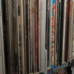 May include: A collection of vinyl records, showcasing album covers with various colors and designs. Titles and artist names are visible, including albums by The Beatles, Bob Dylan, and Miles Davis. The records are stored vertically, suggesting a music collection.