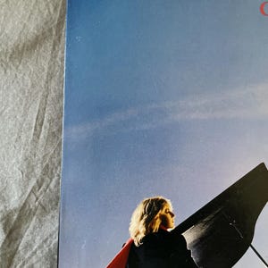 May include: Album cover featuring a woman with blonde hair wearing a black coat and a red scarf, set against a blue sky. A grand piano is in the background, with the woman's silhouette visible.