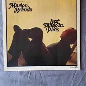 May include: Vintage vinyl record album cover for "Last Tango in Paris" featuring Marlon Brando. The cover has a sepia tone with the film title and credits in stylized fonts. The image shows a silhouette of a person reclining.