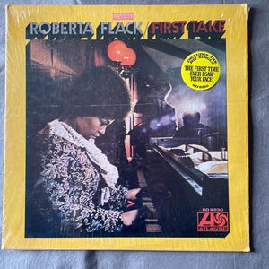 May include: A yellow vinyl record album cover with the title "Roberta Flack First Take" in black text. The cover features a photo of Roberta Flack playing a piano. The album includes the hit single "The First Time Ever I Saw Your Face".