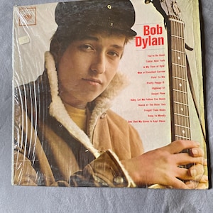 May include: Vintage Bob Dylan album cover featuring a portrait of the musician in a brown jacket and black cap, holding a guitar. The album title and song list are visible on the cover.